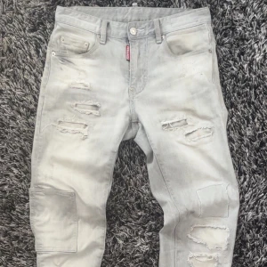 Grå ripped jeans från Dsquared2 - Snygga grå jeans från Dsquared2 med slitna detaljer och trasiga partier på både fram- och baksida. Modellen har en smal passform, klassiska fem fickor och en extra ficka på benet. Jeansen är tillverkade i bomull och har metallknappar med Dsquared2-logga.
