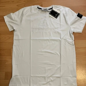 Vit Stone Island t-shirt i bomull - Vit t-shirt från Stone Island med broderad logga på bröstet och klassisk patch på ärmen. T-shirten är i mjuk bomull och har en normal passform med rund hals och korta ärmar. Perfekt för dig som gillar stilrena och exklusiva plagg.
