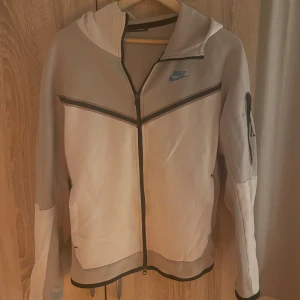 Nike Tech Fleece hoodie beige - Nike Tech Fleece hoodie i beige och grått med svarta detaljer och blå Nike-logga på bröstet. Jackan har hel dragkedja, huva och en ärmficka med dragkedja. Snygg sportig look och skön passform, perfekt för chill eller träning.