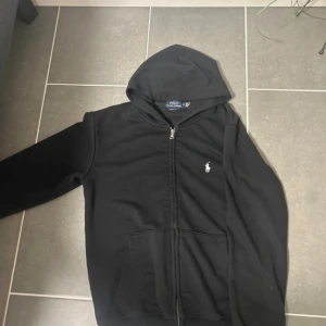 Svart hoodie från Polo Ralph Lauren - Svart hoodie från Polo Ralph Lauren med dragkedja och huva. Tröjan har klassisk logga broderad på bröstet, två fickor fram och är tillverkad i mjuk bomull. Custom fit-modell som sitter snyggt och bekvämt. Perfekt för chill dagar eller när du vill ha en clean look.