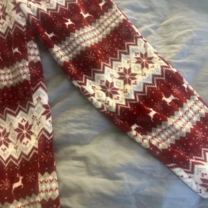 Röda byxor med julmönster - Mysiga röda byxor med vitt och vinrött julmönster, snöflingor och renar. Byxorna har elastisk midja och är perfekta för chill hemma under vintern. Materialet är mjukt och värmande, med klassiskt säsongsmotiv. Snöret borta men det är resår så funkar bra ändå