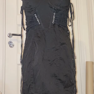 Svart kort klänning med snörning - Svart kort klänning med cool snörningsdetalj längs sidorna och framtill, samt dragkedja i ryggen. Klänningen har en figurnära passform och bustier-inspirerad överdel. Perfekt för dig som gillar edgy och trendiga plagg.