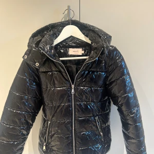 Svart pufferjacka från Nelly - Snygg svart pufferjacka från Nelly i glansigt material med huva och dragkedja framtill. Jackan har två fickor med dragkedja och quiltad design som ger en cool look. Perfekt för dig som vill ha en varm och trendig jacka med lite extra attityd.