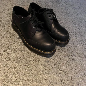 Svarta Dr. Martens skor med snörning - Säljer ett par klassiska svarta Dr. Martens med rund tå och snörning. Skorna har den ikoniska gula sömmen runt sulan och är tillverkade i slitstarkt skinn. Perfekt för dig som gillar chunky och tidlös stil.