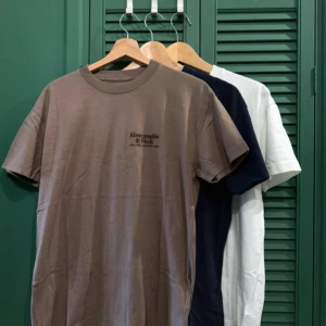 3 pack Abercrombie & Fitch t-shirt i tre färger - Snygga t-shirts från Abercrombie & Fitch i beige, marinblå och vit. Klassisk rund hals och korta ärmar med diskret logotyp på bröstet. Perfekt basic-plagg med stilren look, tillverkad i mjuk bomull för skön känsla hela dagen.
