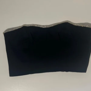 Svart tubtopp med paljetter - Snygg svart tubtopp med glittrande strass längs överkanten. Toppen är axelbandslös och har en stilren bandeau-form som ger en cool och festlig vibe. Perfekt för dig som vill sticka ut med lite extra bling. Endast använd en gång. 