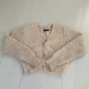 Kofta XS Nyskick, Soaked in Luxury, Smyckesknappar - Koftan har vackra och glittriga smyckesknappar i form av blommor som ger den en lyxig och elegant touch. Knapparna gör koftan perfekt för både vardag och fest. Passform: Koftan är en kortare modell med V-ringning och slutar ungefär vid byxlinningen/midjan. * Ärmar: Långa ärmar med bred mudd.  * Färg: Ljus neutral färg, en blandning av ljusbeige/off white och en svag gråmelerad ton (ofta kallad Oatmeal) * Material:  59% Bomull, 17% Ull, 14% Polyester & 10% Alpaca.