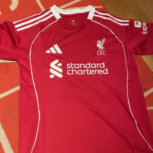 Liverpool FC röd matchtröja Adidas S - Liverpool FCs officiella matchtröja i rött från Adidas, slim fit. Klassisk design med vita detaljer, broderat klubbmärke och sponsortryck på bröstet. Tillverkad i lätt och ventilerande polyester, perfekt för fotboll eller supporterliv.