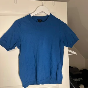 Blå stickad topp från Lindex - En enkel och stilren blå stickad topp från Lindex med rund halsringning och korta ärmar. Toppen har ribbade kanter vid hals, ärmslut och nederkant. Perfekt för dig som gillar en klassisk look med en pop av färg.