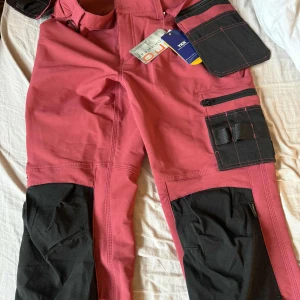 Rosa arbetsbyxor Pro Workwear - Snygga rosa arbetsbyxor från Pro Workwear Bluewear med svarta förstärkningar på knän och fickor. Byxorna har flera praktiska fickor, hällor och detaljer för verktyg. Materialet är slitstarkt och passar dig som vill ha funktion och stil på jobbet.