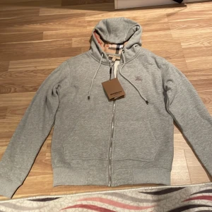 Grå hoodie från Burberry med dragkedja - Säljer en grå hoodie från Burberry med klassiskt rutigt foder i beige, svart och rött. Tröjan har huva med snörning, dragkedja framtill och två fickor. Perfekt för dig som gillar stilrena detaljer och ikoniskt mönster.