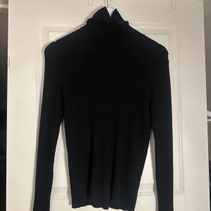 Svart ribbad polotröja från Indiska - Svart polotröja från Indiska i storlek S. Tröjan är ribbstickad med lång ärm och har en klassisk hög krage. Materialet är mjukt och stretchigt, perfekt för lager-på-lager. Enkel och stilren design som funkar till många olika looks.