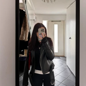 Svart skinnjacka med bälte - Säljer en svart skinnjacka med coola detaljer som bälte i midjan och sned dragkedja. Jackan har klassisk bikerstil med långa ärmar och stilren look, perfekt för dig som vill ha en edgy vibe. Passar till många olika outfits och ger en snygg touch.