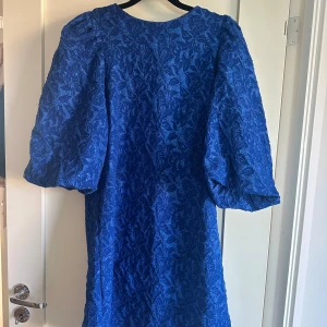 Blå klänning med puffärmar - Säljer en blå klänning med vackert blommigt mönster och puffiga, korta ärmar. Klänningen har en rak siluett och är tillverkad i ett strukturerat tyg som ger en lyxig känsla. Perfekt för dig som vill sticka ut med färg och volym. Går ner till knäna.