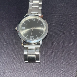Stilren svart klocka med metallarmband - Säljer en stilren klocka med svart urtavla och romerska siffror. Klockan har ett klassiskt runt hölje i silverfärgad metall och matchande länkarmband. Perfekt accessoar för dig som gillar en clean och tidlös look.