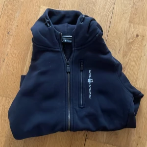 Svart väst från Sail Racing med huva - Svart väst från Sail Racing med huva och dragkedja framtill. Västen har en extra bröstficka med dragkedja och logga broderad på bröstet. Materialet är mjukt och känns som syntet, perfekt för lager-på-lager. Snygg och sportig look.