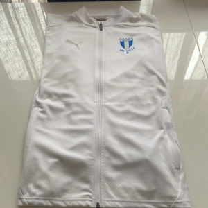 Vit jacka från Puma med Malmö FF-logga - Säljer en stilren vit jacka från Puma med Malmö FF-logga på bröstet. Jackan har dragkedja framtill och är långärmad. Perfekt för fotbollsfans som vill visa sitt stöd. 🩵