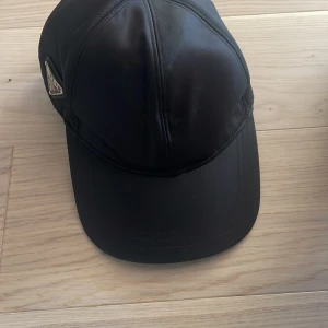 Svart keps från Prada i syntet - Snygg svart keps från Prada med klassisk böjd skärm och justerbar rem baktill. Kepsen har ett stilrent utseende med Pradas ikoniska metallmärke på sidan. Tillverkad i slät syntetmaterial som ger en modern vibe.