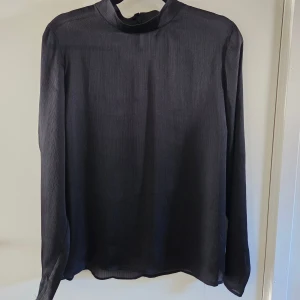 Svart långärmad blus Gina Tricot - Stilren svart blus från Gina Tricot med diskret ribbad struktur och hög hals. Långärmad modell i lätt, glansigt tyg som ger en snygg och elegant look. Stängs med knapp i nacken och har en rak passform.