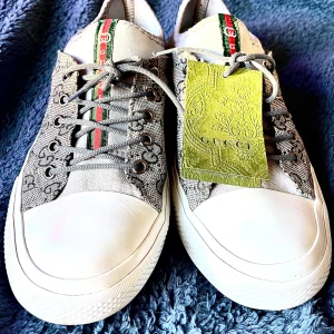 Grå sneakers med GG-mönster  - Säljer ett par grå sneakers som är nästan ny GG-mönster i svart på canvas. Skorna har vita gummisulor, rund tå och snörning. Detaljer med grönt och rött band längs plösen och bak, samt original dustbag och tagg medföljer. Perfekt för dig som gillar lyxiga streetwear-vibes.