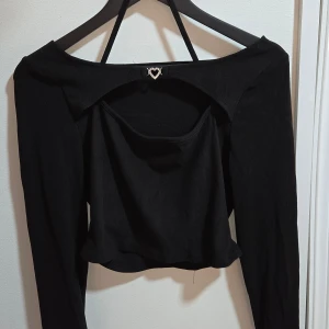 Svart croppad långärmad bolerotopp med strass-hjärta, M - Svart bolerotopp med croppad passform och lång ärm. Toppen har en snygg öppen rygg med dekorativt hjärtdetalj framtill och ett smalt band som går runt halsen. Materialet är mjukt och stretchigt, perfekt för att skapa en cool och trendig look.