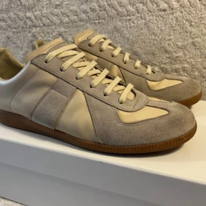 Maison Margiela Gats - Maison Margiela Gats i beige och grå toner och platt gummisula. Ovandelen är i en mix av mocka och skinn, med snörning framtill. Snygg retrostil som passar till jeans eller dressade byxor. Diskret logga på sulan och stilrena detaljer. Använda ca 5 gånger i somras, säljs för 2999kr och är i storlek 42. 