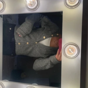 Grå stickad kofta med volangdetalj - Mysig grå stickad kofta med snygga guldknappar och volangdetalj över axlar och rygg. långa ärmar, perfekt för lager på lager. Passar dig som gillar en chill men ändå söt stil.