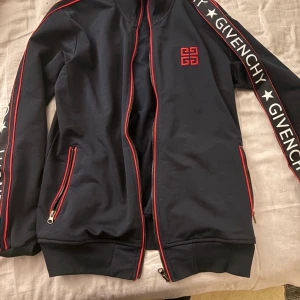 Svart Givenchy bomberjacka med röda detaljer - Svart bomberjacka från Givenchy med röda detaljer och dragkedjor. Jackan har logga på bröstet och breda band med Givenchy-tryck längs ärmarna. Snygg och sportig look med ribbade muddar och hel dragkedja framtill.