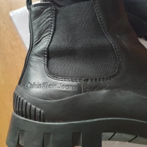Svarta chelsea boots från Calvin Klein - Snygga svarta chelsea boots från Calvin Klein Jeans med grov sula och elastiska paneler på sidorna. Skorna är i skinn och har diskret logga på sidan samt bak på sulan. Perfekta för dig som vill ha en stilren och robust look.
