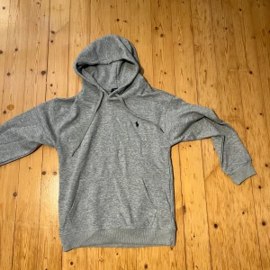 Grå hoodie från Polo Ralph Lauren - Säljer en grå hoodie från Polo Ralph Lauren i storlek S. Tröjan har huva med snörning, klassisk logga broderad på bröstet och ribbade muddar vid ärmslut och nederkant. Perfekt för chill dagar och enkel att matcha med jeans eller joggers.