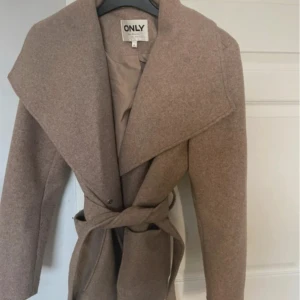 Beige kappa från ONLY med bälte - Stilren beige kappa från ONLY med bred krage och knytskärp i midjan. Jackan har långa ärmar, två fickor framtill och är tillverkad i ett ull-liknande material. Perfekt för dig som vill ha en klassisk och snygg look. Den är lite nopprig på ena armen syns knappt, inte använd så mycket