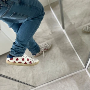 Saint Laurent sneakers med röda stjärnor - Snygga vita sneakers från Saint Laurent med röda stjärnor på sidorna och blå detalj vid hälen. Skorna har snörning och platt sula, tillverkade i skinn med coolt mönster. Perfekta för dig som gillar att sticka ut med din stil. 