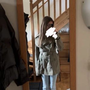 Grön Jacka från vera Moda  - dubbelknäppning och bälte i midjan. Klassisk krage och axelklaffar ger en stilren look. Perfekt för dig som vill ha en tidlös jacka med coola detalje