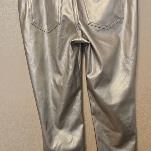 Silvriga syntetbyxor L/XL - Snygga byxor i metallic silver med klassisk femficksdesign och bälteshällor. Materialet är syntet med skinnliknande finish som ger en cool och modern vibe. Passformen är normal och midjan är midwaist. Perfekt för dig som vill sticka ut med en unik look.