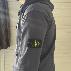 Mörkgrå hoodie från Stone Island - Säljer en mörkgrå hoodie från Stone Island med ikonisk patch på ärmen. Tröjan har huva med snörning, dragkedja framtill och ribbade muddar. Tillverkad i mjuk bomull, perfekt för chill dagar. Snygg och enkel design som alltid funkar.