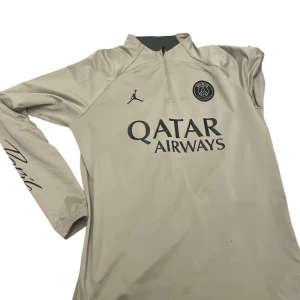 Beige PSG långärmad fotbollströja - Snygg beige långärmad fotbollströja från PSG och Jordan med halv dragkedja, svart Paris Saint-Germain-logga och Qatar Airways-tryck på bröstet 