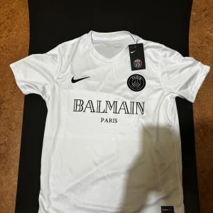 Vit PSG x Balmain fotbollströja Nike - Snygg vit fotbollströja från Nike i samarbete med PSG och Balmain. Tröjan har korta ärmar, rund halsringning och svart PSG-logga samt Balmain Paris-tryck på bröstet. Tillverkad i lätt och glansigt funktionsmaterial som andas.