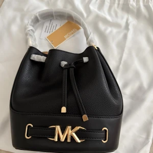 Svart handväska från Michael Kors - Snygg svart handväska från Michael Kors med guldfärgade MK-detaljer framtill. Väskan har en rundad form, snörning upptill och är tillverkad i skinn med både slät och strukturerad yta. Handtaget är vadderat och insidan är rymlig för dina essentials.