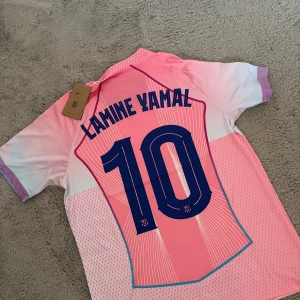 Barça Lamine Yamal rosa matchtröja Nike - Säljer en FC Barcelona fotbollströja med Lamine Yamal och nummer 10. Tröjan är rosa med lila och vita detaljer, har korta ärmar, V-ringning och tryckta loggor från Nike, Spotify och UEFA Foundation. Materialet är lätt och andas, perfekt för match eller träning.