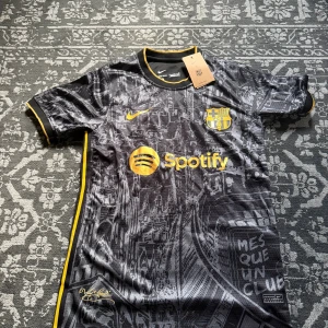 Barcelona svart matchtröja Nike - Snygg FC Barcelona fotbollströja från Nike i svart med detaljer i gult. Tröjan har korta ärmar, rund hals och coolt grafiskt mönster med stadsmotiv. Officiella klubbmärket, Spotify-logga och texten 'Mes Que Un Club' på framsidan. Materialet är lätt och andas.