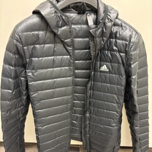 Grå pufferjacka från Adidas med huva - Snygg grå pufferjacka från Adidas med huva och diskret logga på bröstet. Jackan har horisontella quiltade paneler och dragkedja framtill. Perfekt för kyliga dagar och har en modern, sportig look som funkar till streetwear.