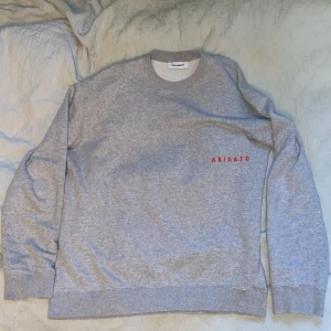 Grå sweatshirt från Axel Arigato - Säljer en stilren grå sweatshirt från Axel Arigato med röd broderad text på bröstet. Tröjan har rund halsringning, långa ärmar och är tillverkad i mjuk bomull. Perfekt för en clean och avslappnad look.
