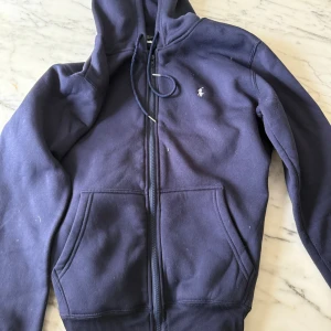 Marinblå hoodie från Ralph Lauren - Säljer en marinblå hoodie från Ralph Lauren med dragkedja och huva. Tröjan har klassisk broderad logga på bröstet, kängurufickor och ribbade muddar. Perfekt för en chill och stilren look.