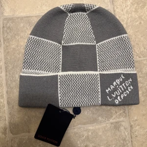 Louis Vuitton grå rutig mössa - Snygg grå och vit mössa från Louis Vuitton med rutigt mönster och broderad text på sidan. Tillverkad i lyxig blandning av kashmir, ull och silke. Perfekt accessoar för dig som vill sticka ut med designerstil och mjukt material.