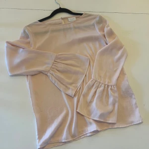 Ljusrosa blus med trumpetärmar från Vila - Supersöt ljusrosa blus från Vila med vida trumpetärmar och rund halsringning. Blusen har en knapp i nacken och är tillverkad i ett mjukt, glansigt material som ger en lyxig känsla. Perfekt för dig som gillar romantiska och feminina detaljer.