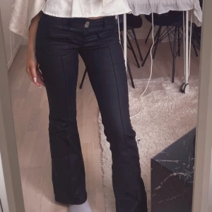 Svarta skinnbyxor bootcut nelly - Säljer ett par svarta bootcut byxor med snygga fickdetaljer framtill och markerad söm längs benen. Byxorna har låg midja och sitter tajt upptill med utsvängda ben. Perfekta för dig som gillar en trendig och stilren look.