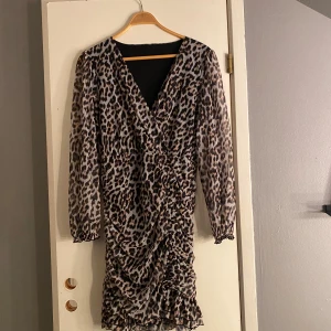 Leopardmönstrad omlottklänning - Snygg kort leopardmönstrad klänning med omlott och v-ringad front. Klänningen har långa, transparenta ärmar med volangdetalj vid ärmslut och rynkade detaljer längs kjolen. Materialet är lätt och ger en cool vibe, perfekt för dig som gillar att sticka ut.😎🐈‍⬛💛