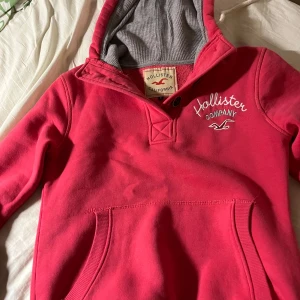 Rosa hoodie från Hollister med huva - Säljer en snygg rosa hoodie från Hollister med broderad logga på bröstet och stor känguruficka framtill. Tröjan har grått innerfoder i huvan och är långärmad. Perfekt för dig som gillar en avslappnad och sportig stil. Accepterar bud!!