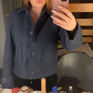 Mörkblå kappa - Snygg mörkblå jacka i ull med klassisk krage och stora knappar framtill. Jackan har två rejäla fickor och raka, långa ärmar med knappdetalj vid ärmslutet. Perfekt för dig som gillar stilrena och tidlösa plagg.