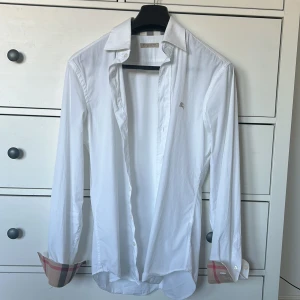 Vit Burberry Brit skjorta med rutiga detaljer - Vit skjorta från Burberry Brit i bomull med klassiska rutiga detaljer på insidan av manschetterna. Skjortan har lång ärm, knappar framtill och liten broderad logga på bröstet. Snygg och stilren modell med ikoniska Burberry-rutor som syns när du viker upp ärmarna.